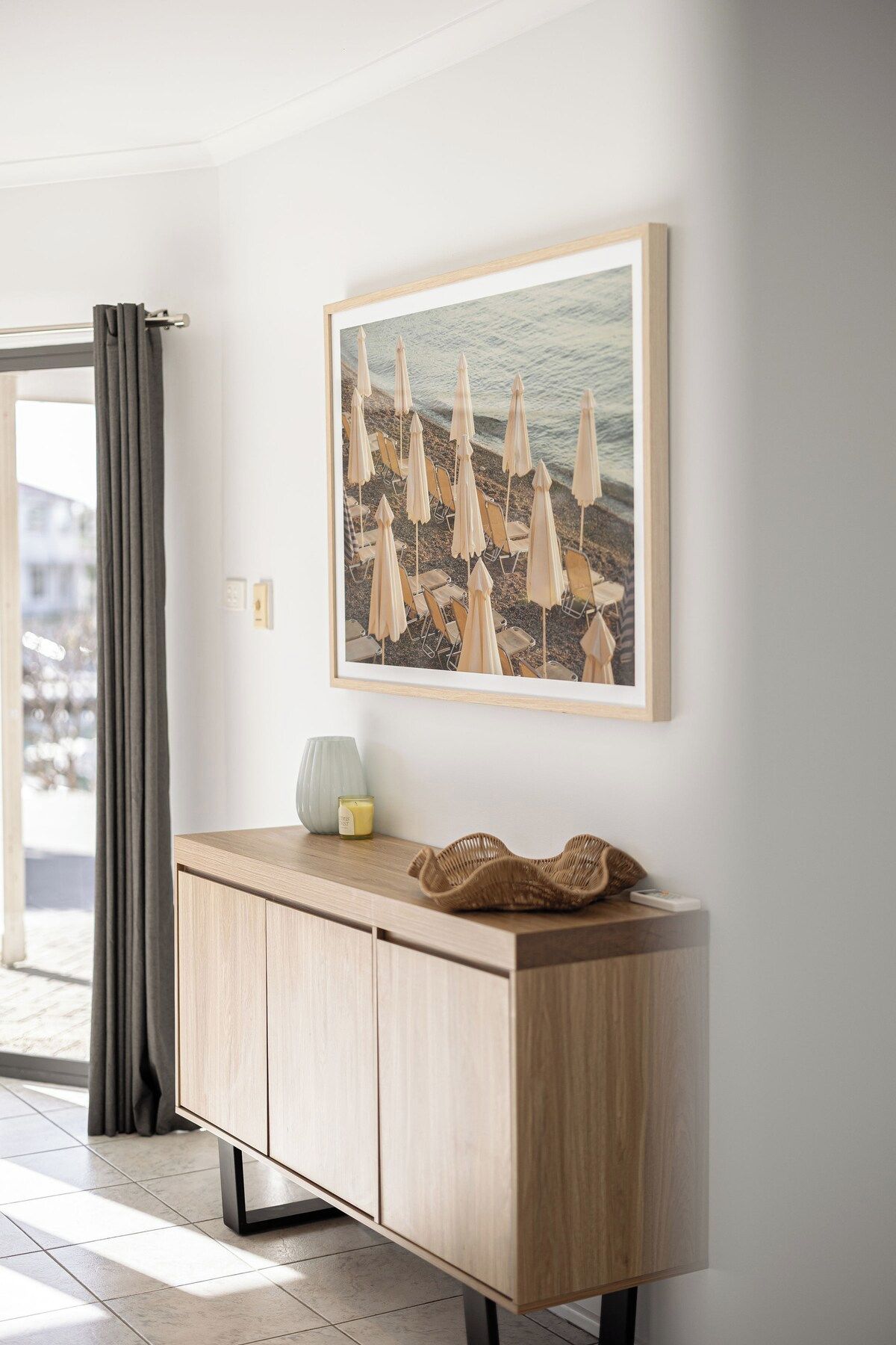 Une pièce avec un meuble en bois clair, un tableau représentant une scène de plage et une fenêtre avec des rideaux.