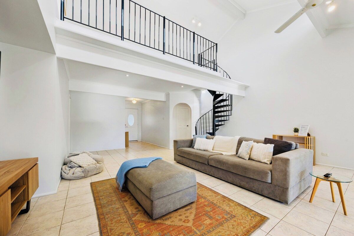 A spacious living room with a high ceiling, a grey sofa set, a wooden coffee table, and a patterned rug.