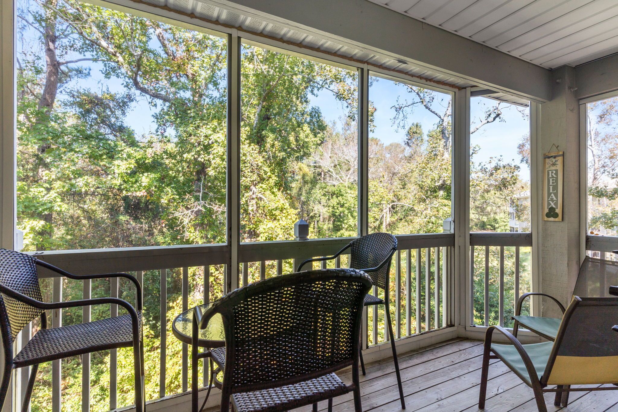 A covered porch with wicker chairs and a table, overlooking a wooded area.