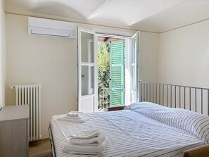 Une chambre à coucher avec un lit, un radiateur, une fenêtre ouverte avec des volets verts, et une vue sur la verdure à l’extérieur.