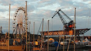Un puerto deportivo con barcos amarrados, una noria y una grúa con la etiqueta 'ROSTOCKER' al fondo.