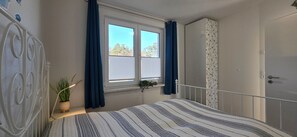 Ein Schlafzimmer mit Bett, einem Fenster mit Vorhängen, einer Topfpflanze und einem Kleiderschrank.