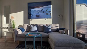 Una sala de estar moderna con un sofá seccional gris, una mesita auxiliar, una mesa de centro redonda y un cuadro grande enmarcado con una escena de playa.