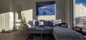 Ein modernes Wohnzimmer mit einem grauen Ecksofa, einem kleinen Beistelltisch, einem runden Couchtisch und einem großen Bilderrahmen mit einer Strandansicht.