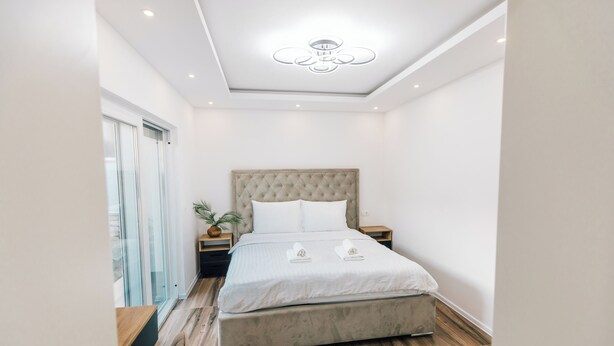 Une chambre à coucher moderne avec un grand lit, une tête de lit capitonnée, des tables de chevet et un luminaire au plafond.