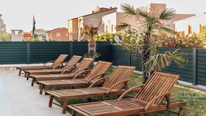 Une rangée de chaises longues en bois est alignée le long d’un chemin de béton, avec un palmier et une clôture verte en arrière-plan.