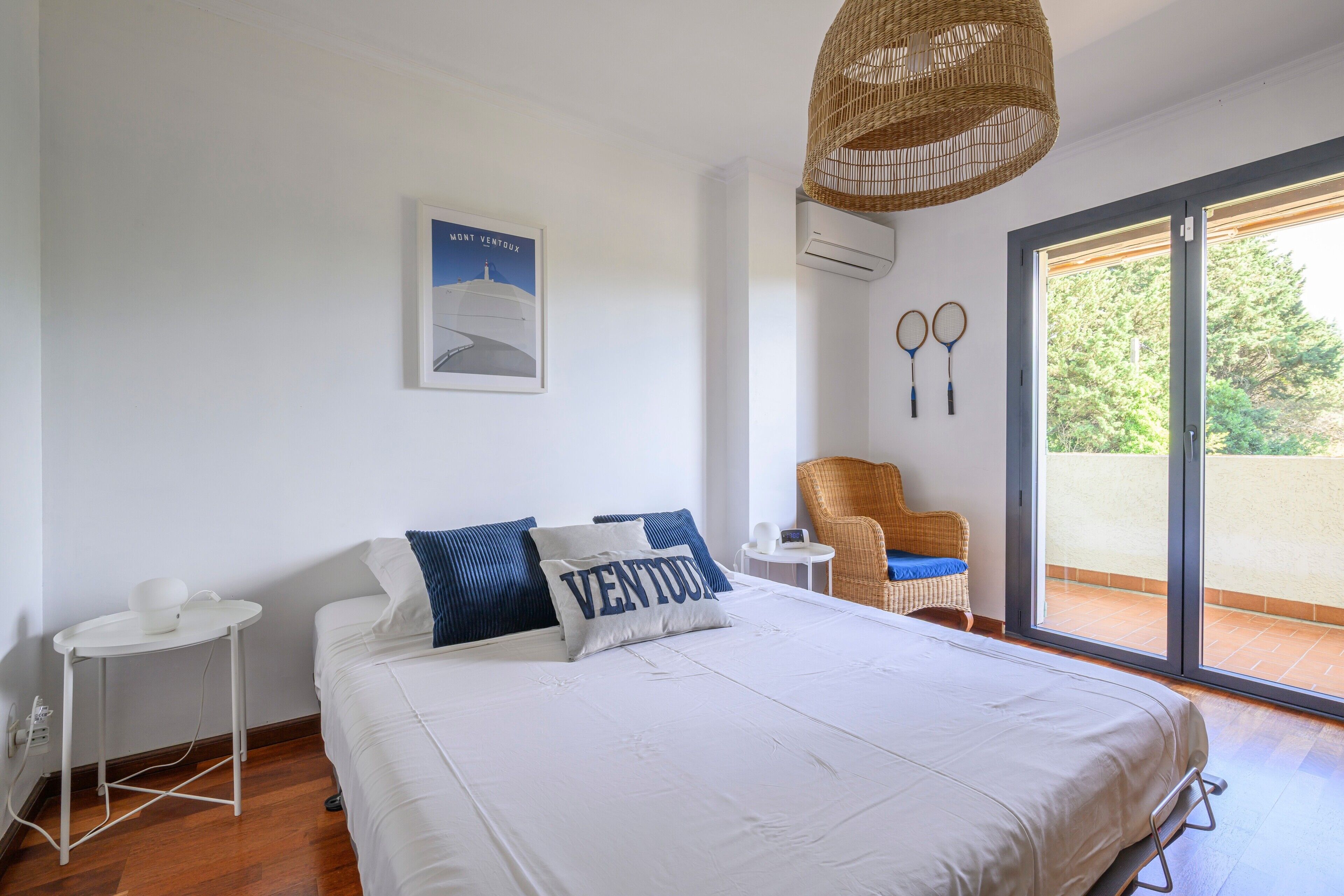 A bedroom with a bed, a wicker chair, a small table, a wall-mounted air conditioner, and a large window with a view.