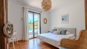 A bedroom with a bed, a wicker chair, a dining table with chairs, a large window, and a ceiling light fixture.