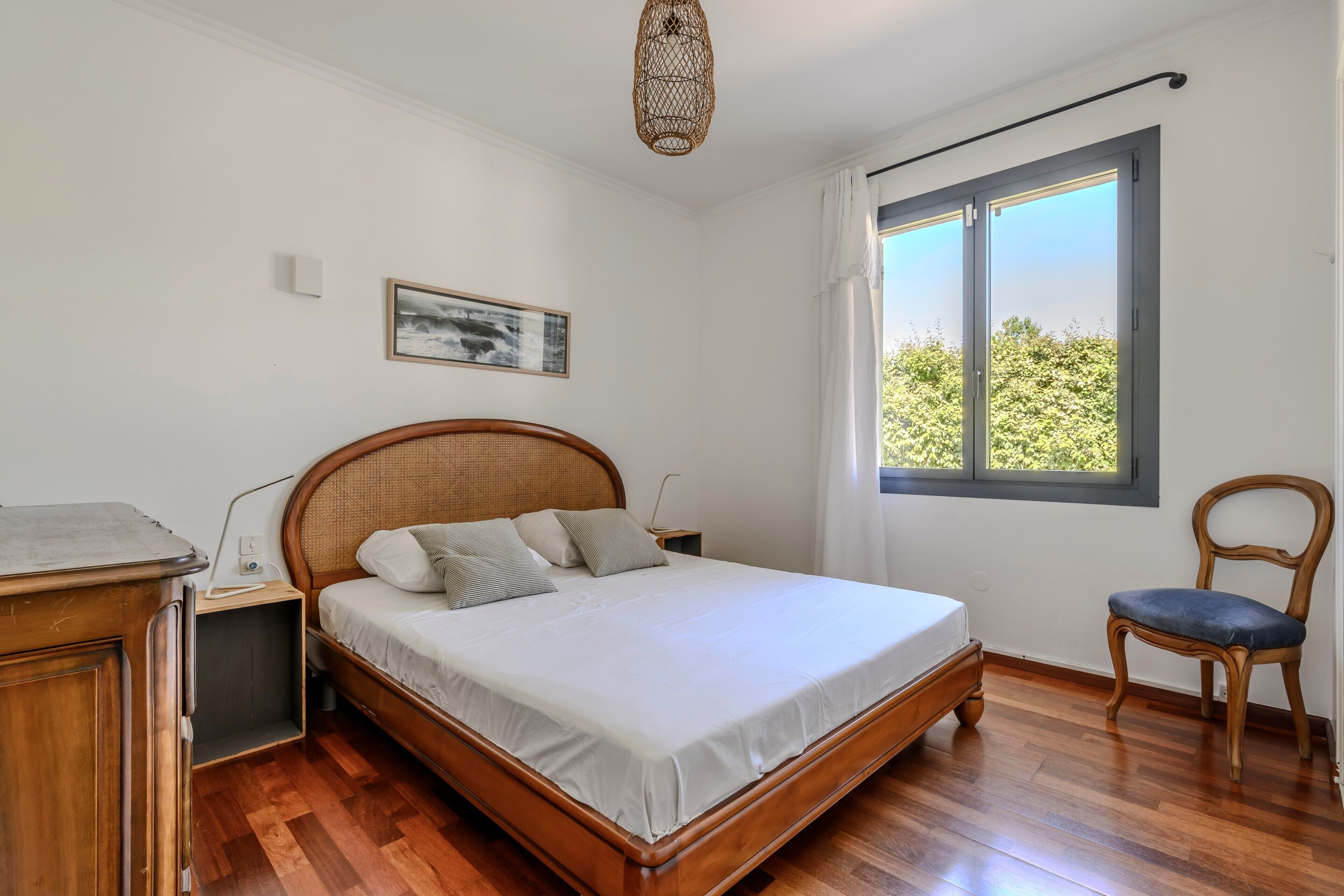 Ein Schlafzimmer mit einem hölzernen Bett, einem Nachttisch, einem Sessel, einem Fenster mit Ausblick und einer Wandleuchte.