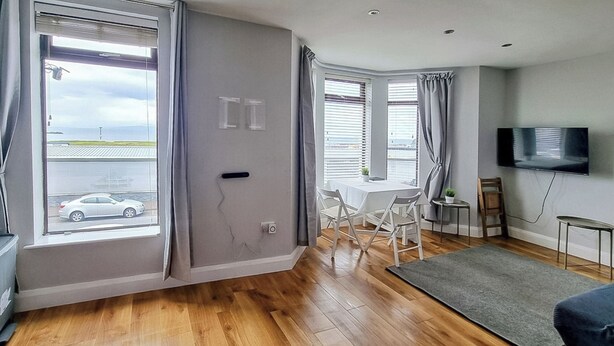 Een moderne woonkamer met houten vloeren, een flatscreen televisie, een eettafel met stoelen en grote ramen met gordijnen.