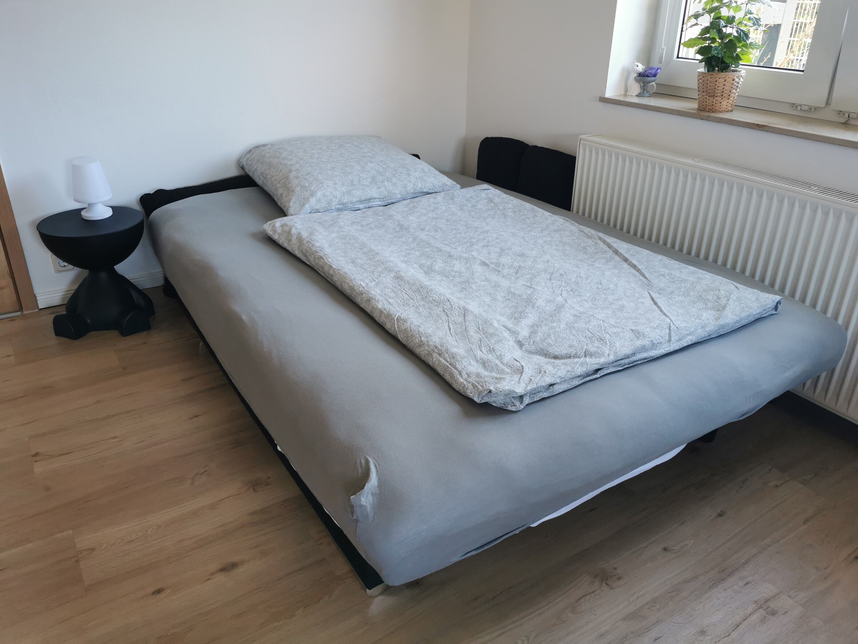 A bed with a grey mattress and a white blanket, a small black side table with a white lamp, and a window with a plant.