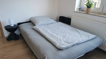 A bed with a grey mattress and a white blanket, a small black side table with a white lamp, and a window with a plant.