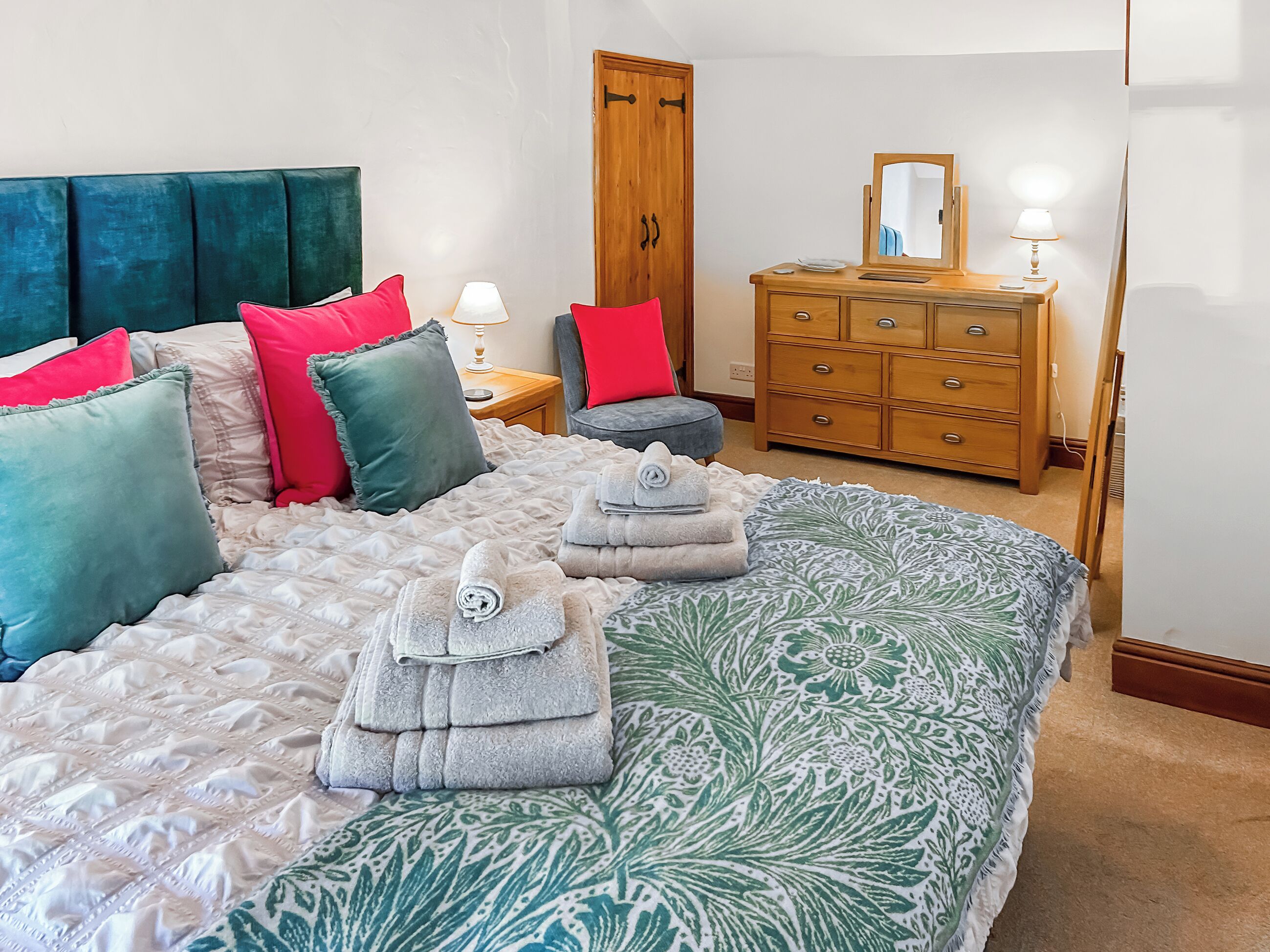 A neatly made bed with multiple pillows and folded towels, a wooden dresser with a mirror, and a bedside table with a lamp.