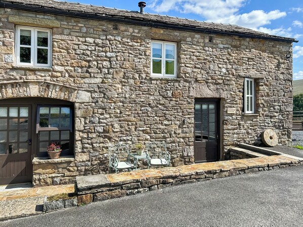 A stone cottage with a front door, two windows, and a small outdoor seating area with a table and chairs.