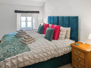 A neatly made bed with a patterned bedspread, multiple pillows in various colors, a stack of folded towels, and a bedside table with a lamp.