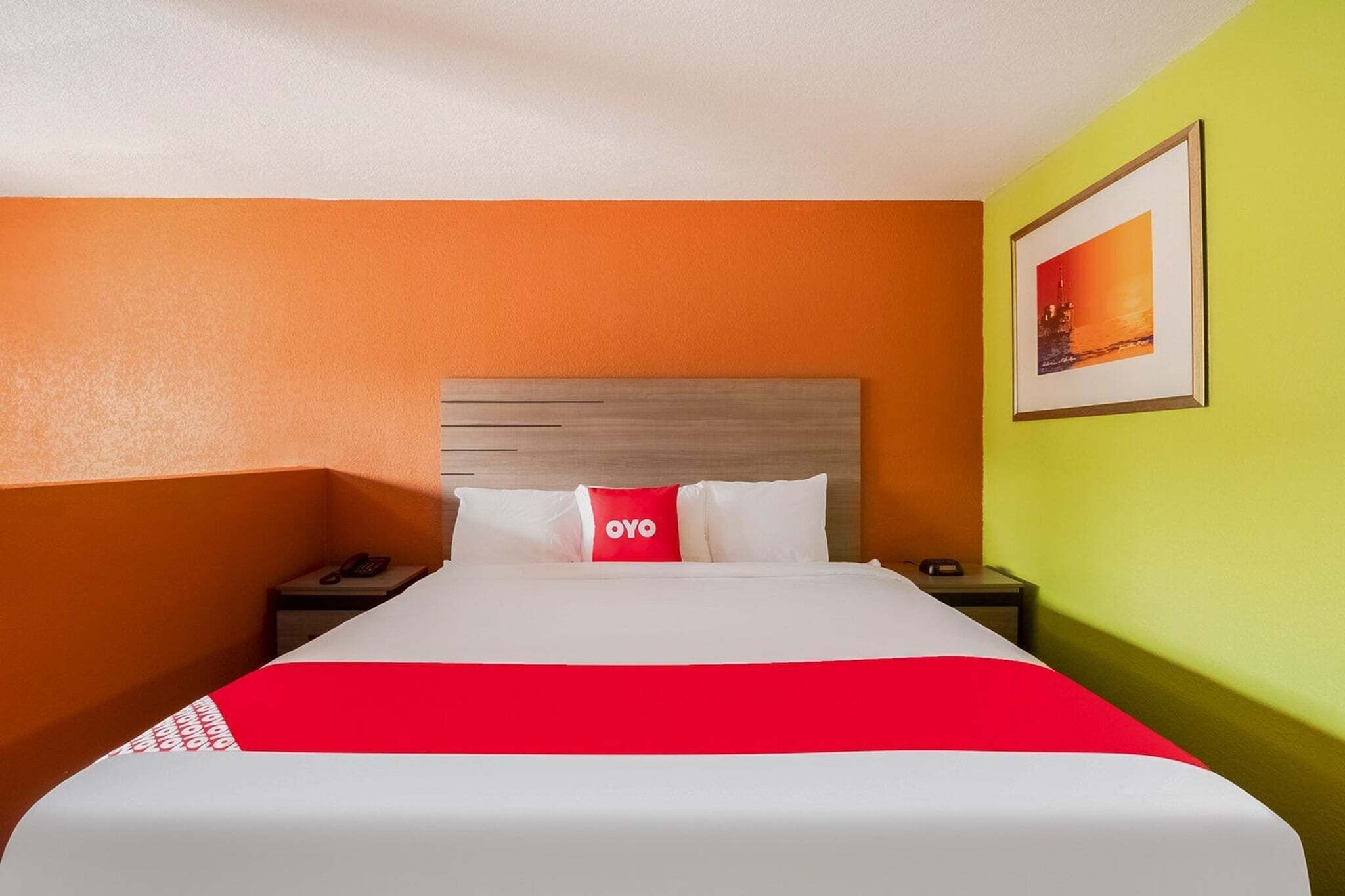 Hotellihuone, jossa on sänky, yöpöytä, punainen tyyny, jossa on teksti »OYO», seinällä oleva kehystetty kuva sekä oranssit ja vihreät seinät.