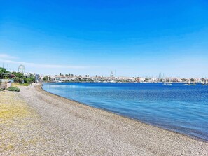 A coastal cityscape with a pebbled beach, a clear blue sky, and a distant view of buildings and a Ferris wheel. - L'île des Marinas by Interhome (Cap d'Agde)