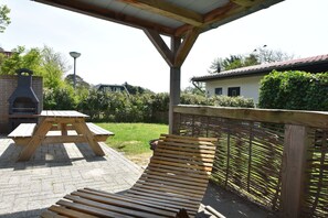Eine überdachte Terrasse mit Holztisch und -bänken, einem Backofen aus Ziegeln und Blick auf ein Haus und Grünflächen.