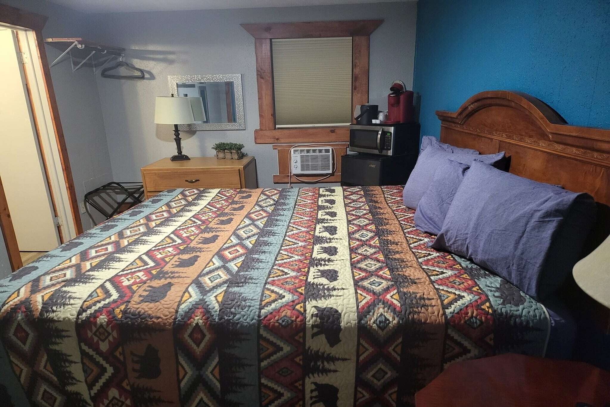A bedroom with a patterned bedspread, a wooden headboard, a dresser, a mirror, and a window with blinds.