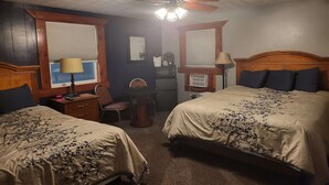 Une chambre d’hôtel avec deux lits, un bureau, une chaise, un ventilateur de plafond et une fenêtre avec des stores.