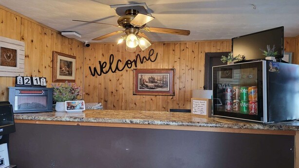A reception area with a wooden wall, a ceiling fan, a welcome sign, a refrigerator, and a coffee machine.