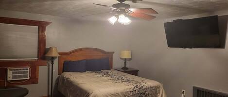 Ein Schlafzimmer mit einem Bett, einem Deckenventilator, einem Fernseher, einem Fenster mit Jalousien, einem Heizkörper und einer Lampe.