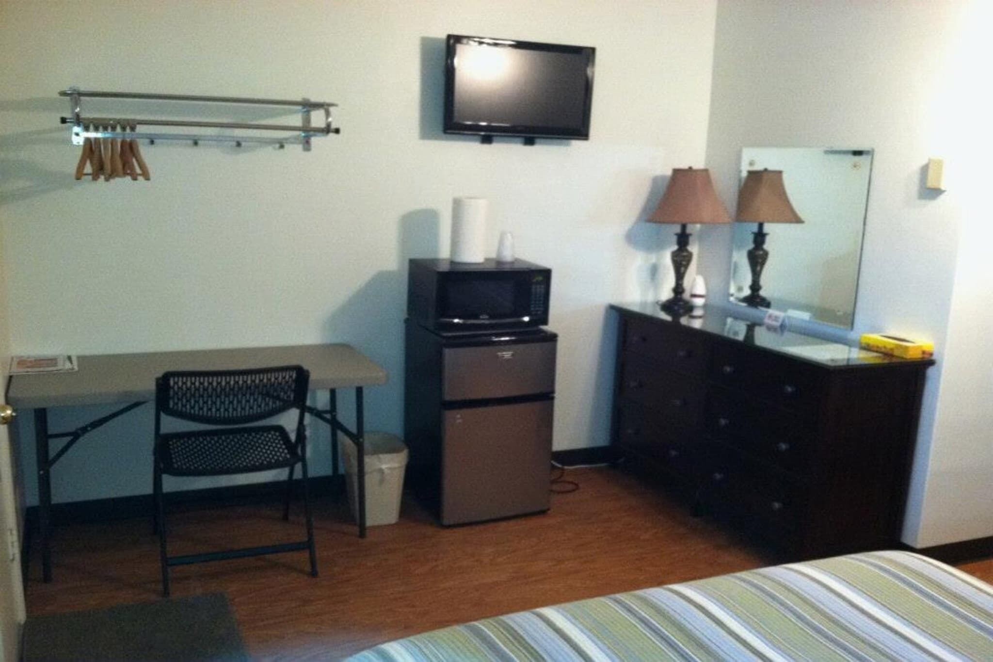 A compact hotel room with a bed, a desk with a chair, a television, a small refrigerator, a dresser, and a mirror.