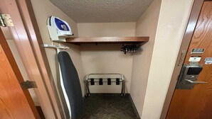 A narrow hallway with a door on the right, a wall-mounted ironing board, a metal rack, and a small shelf with bottles. - Queen Bed Accessible Non-Smoking (Marysville, KS)