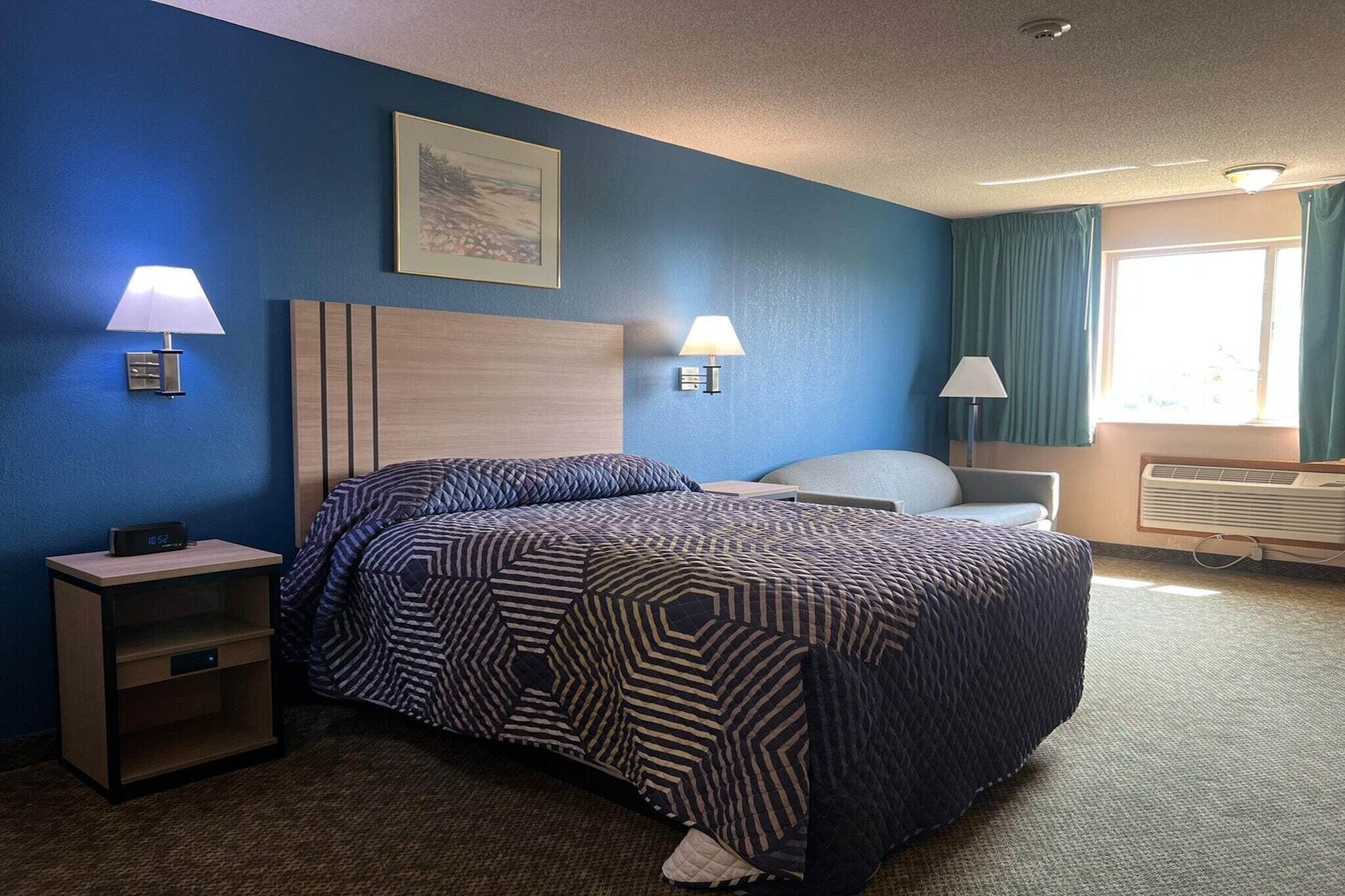 A hotel room with a bed, two bedside lamps, a nightstand, a window with curtains, and a wall-mounted picture.