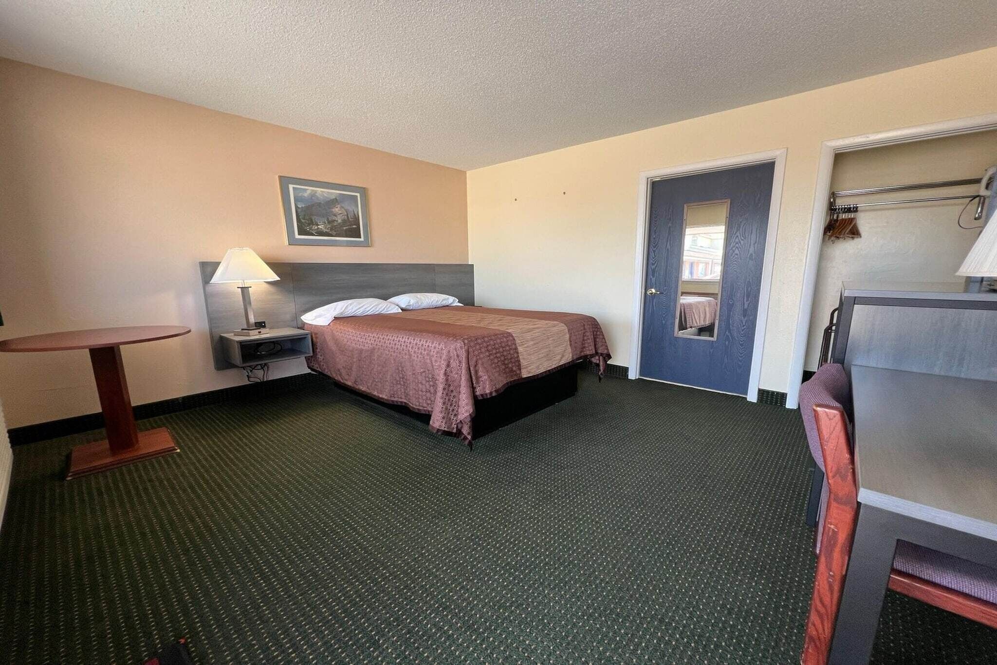 A hotel room with a bed, a desk, a chair, a lamp, a door, and a small table.
