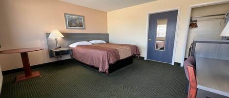 A hotel room with a bed, a desk, a chair, a lamp, a door, and a small table.