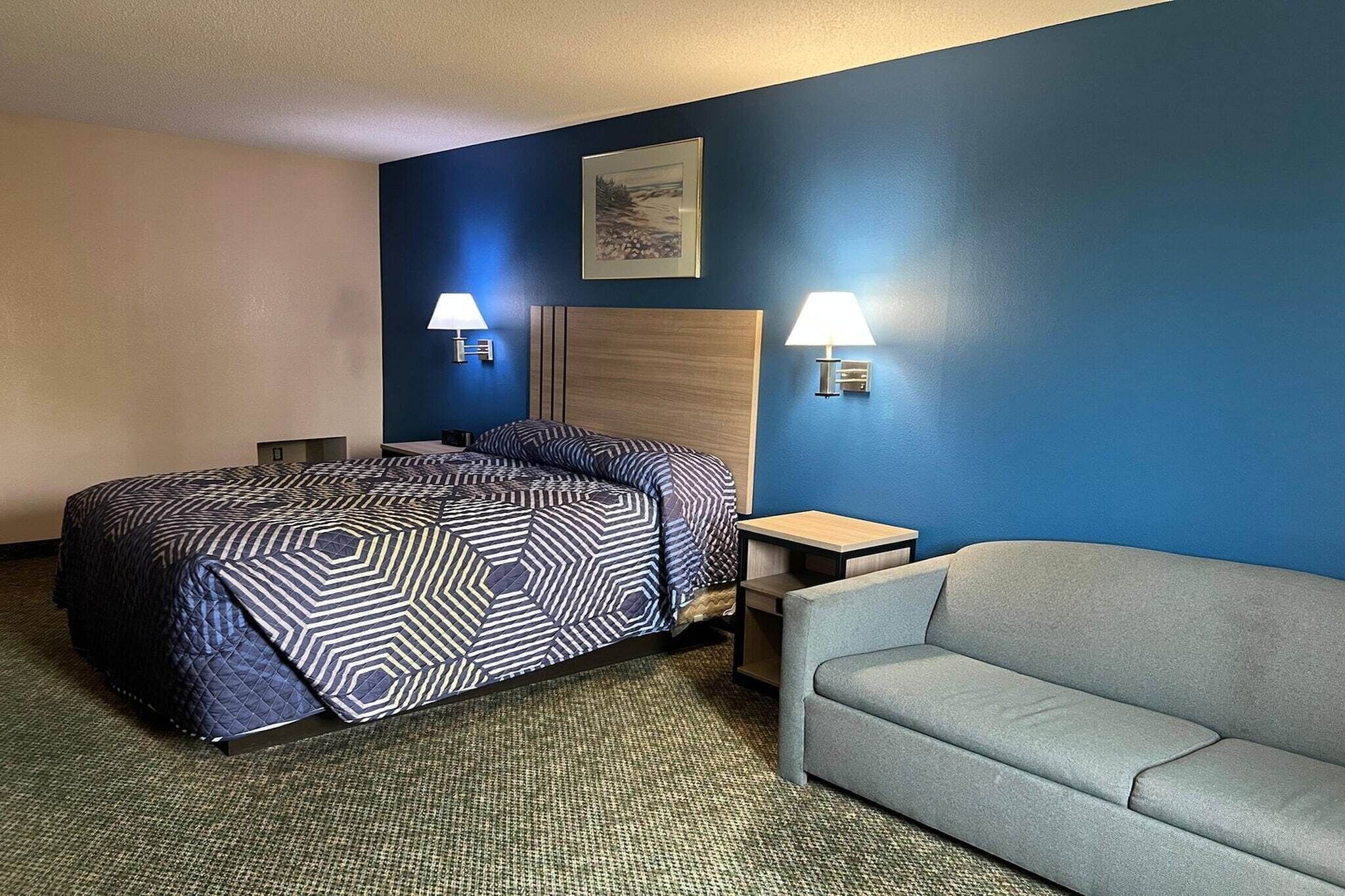 A hotel room with a bed, two bedside lamps, a gray sofa, and a framed picture on the wall.
