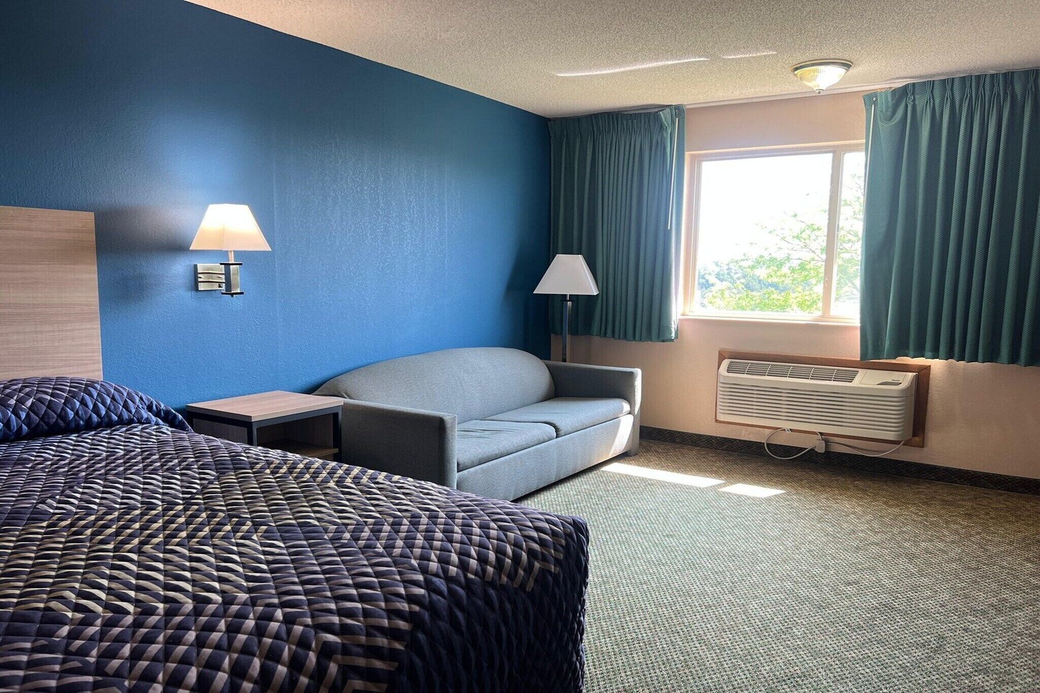 Ein Hotelzimmer mit einem Bett, einem Sofa, zwei Lampen, einem Fenster mit Vorhängen und einer Klimaanlage.