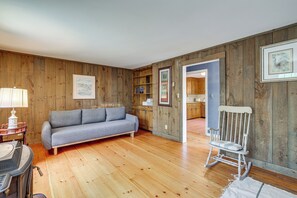 A room with wooden walls, a grey sofa, a rocking chair, and a kitchen visible through an open doorway. - Classic Cape House 5M to Nauset Beach (Orleans)