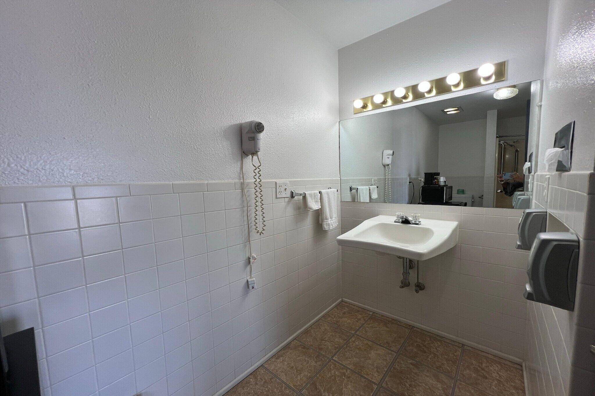 A bathroom with white tiled walls, a sink, a mirror with lights, a hairdryer, and a hand dryer.
