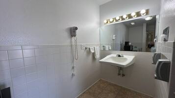 A bathroom with white tiled walls, a sink, a mirror with lights, a hairdryer, and a hand dryer.
