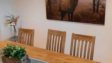 Un coin repas avec une table en bois, trois chaises en bois, un vase de fleurs et un tableau représentant une vache accroché au mur.