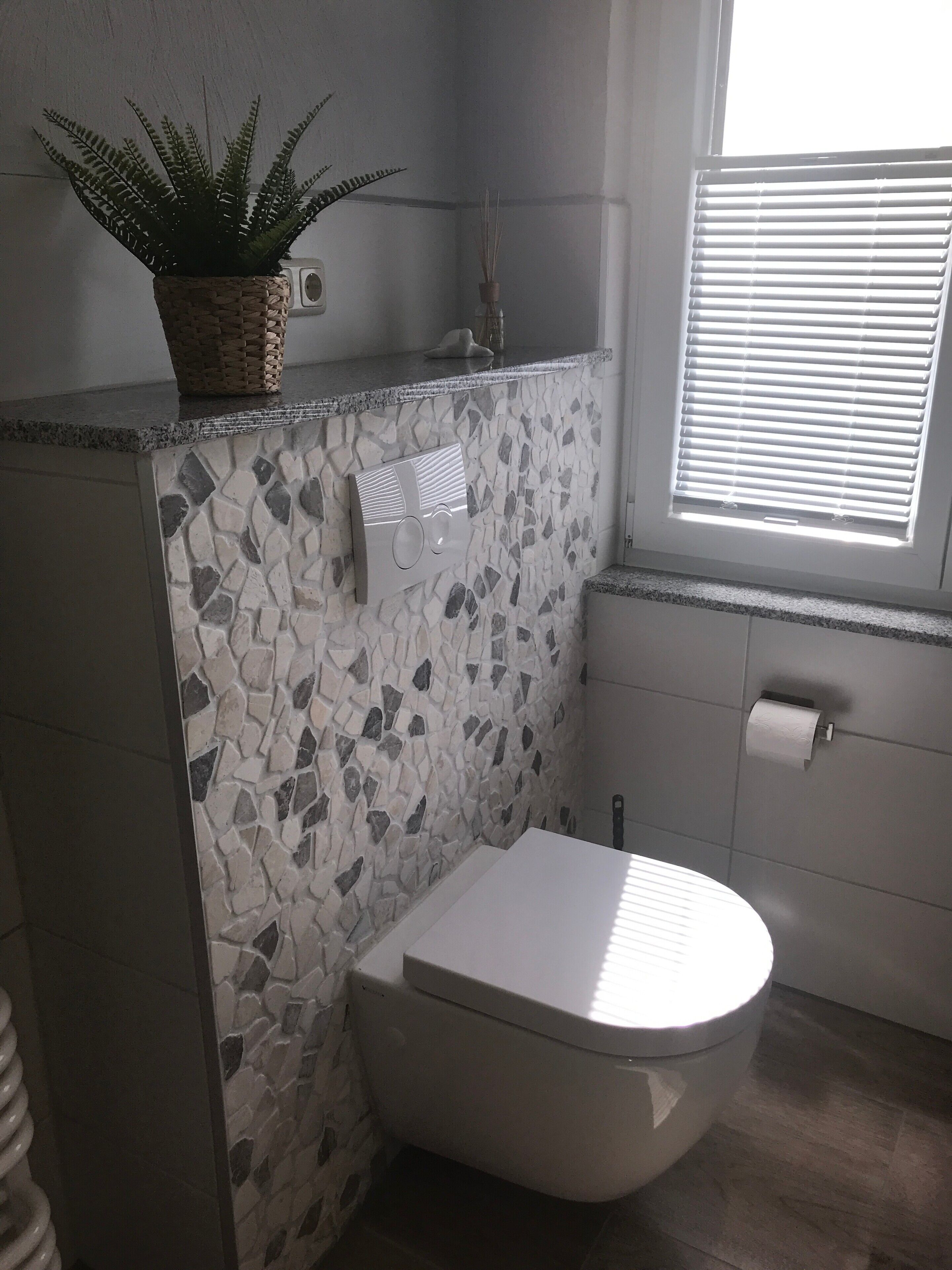A bathroom with a mosaic wall, a toilet, a sink, a window with blinds, and a plant on the counter.
