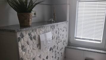 A bathroom with a mosaic wall, a toilet, a sink, a window with blinds, and a plant on the counter.