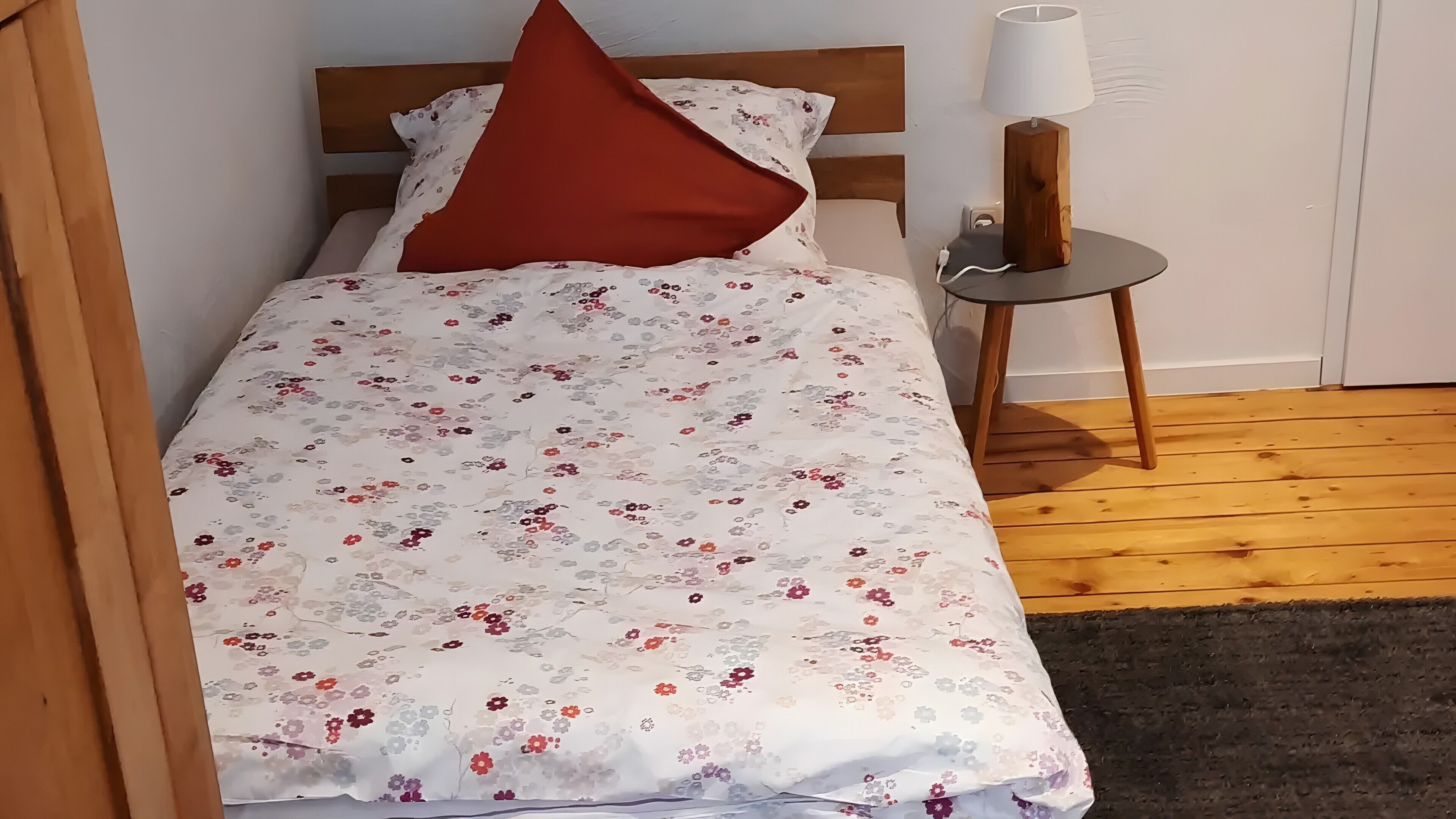A bed with a floral bedspread, a red pillow, and a bedside lamp on a wooden floor.