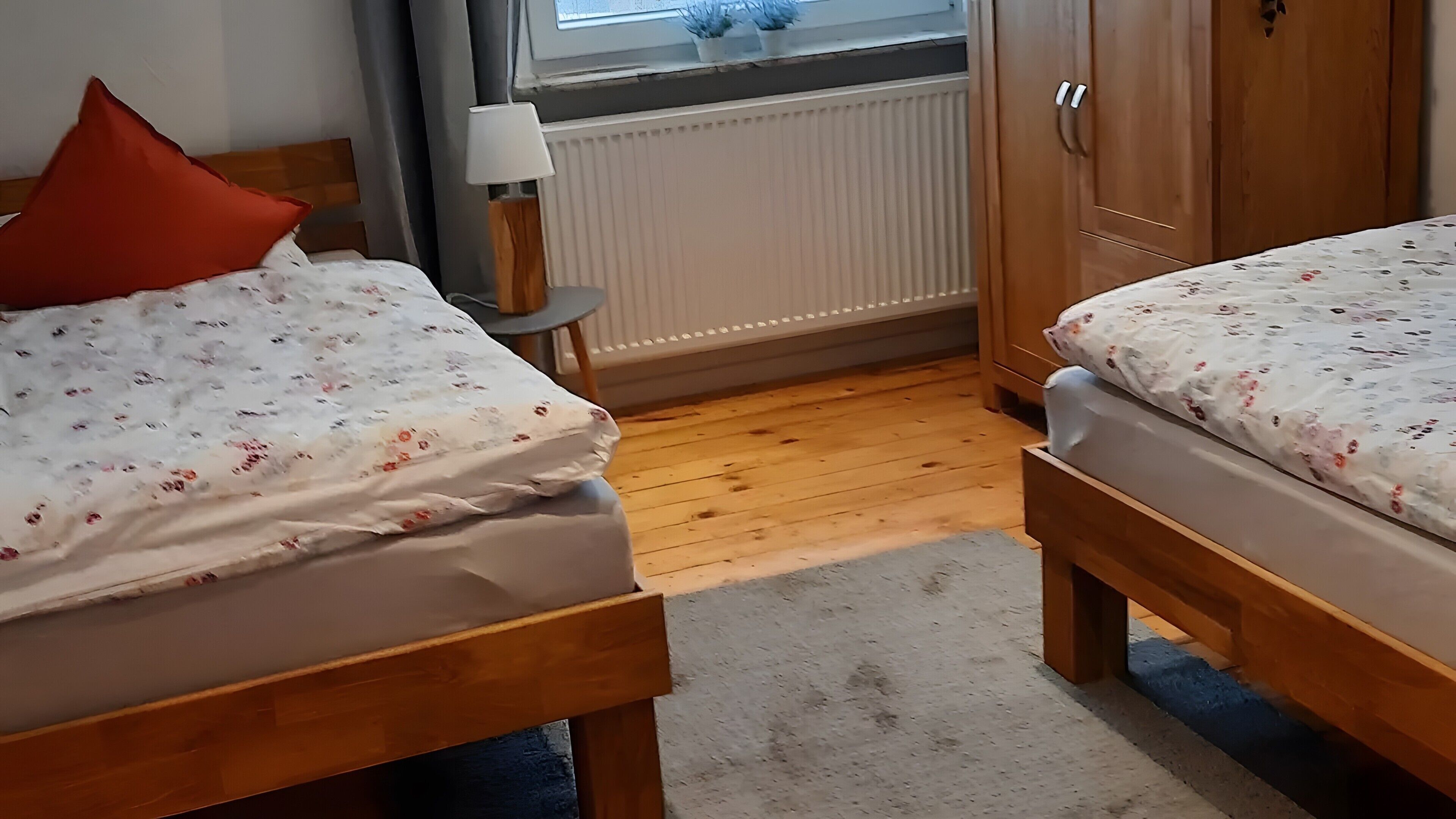 Ett rum med två sängar i trä, ett element, ett fönster med gardiner, ett litet bord med en lampa och en garderob i trä.
