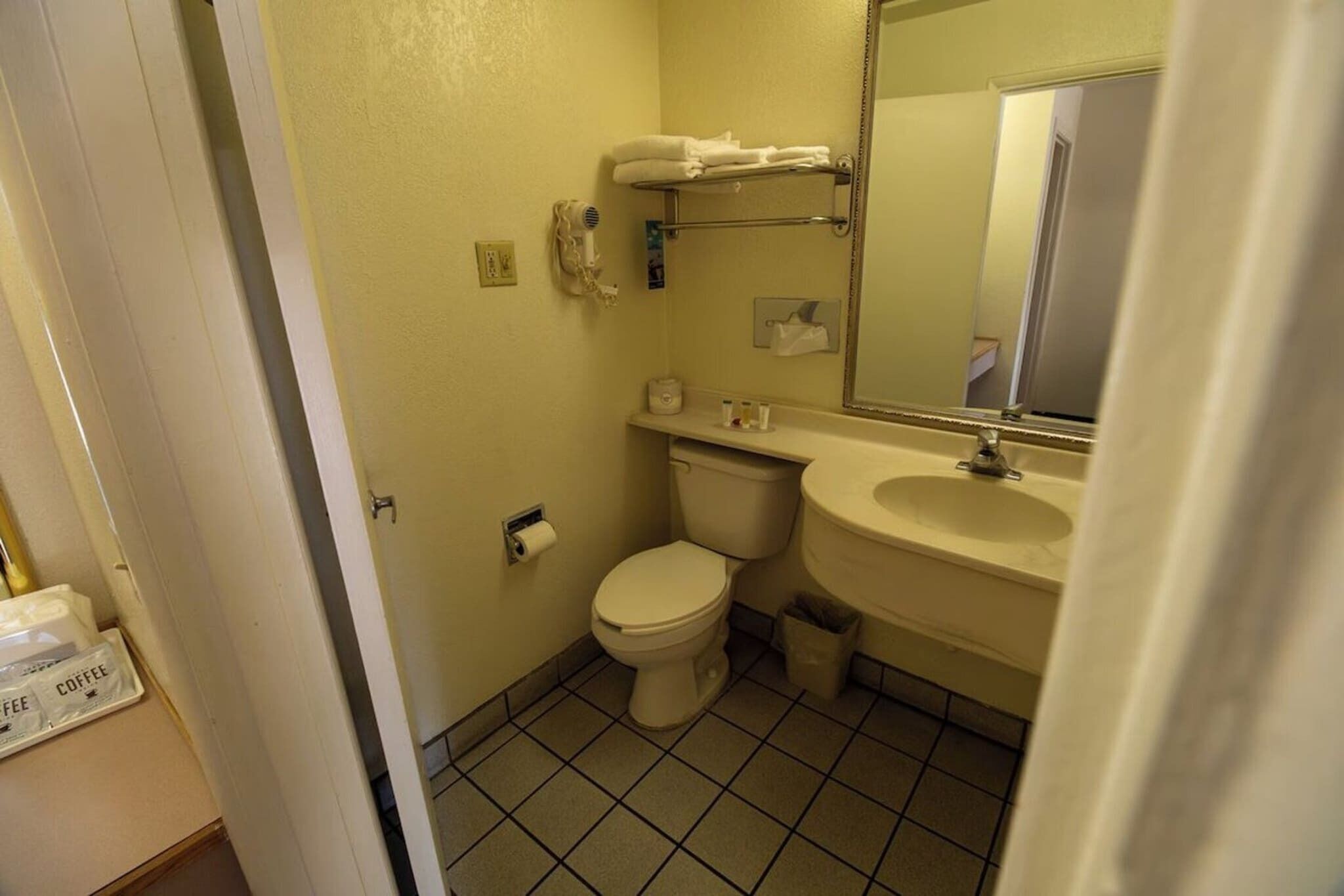 Et lille badeværelse med toilet, håndvask og spejl. Der er en bøjle over vasken og en skraldespand ved siden af toilettet.