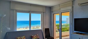 Ein Zimmer mit großem Fenster und Blick auf einen Strand, einem Fernseher auf einem Schrank und einem Sessel.