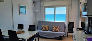 Ein Zimmer mit Esstisch, einer Couch mit gestreiften Kissen, einem großen Fenster mit Meerblick und einem Fernseher auf einem Beistelltisch.