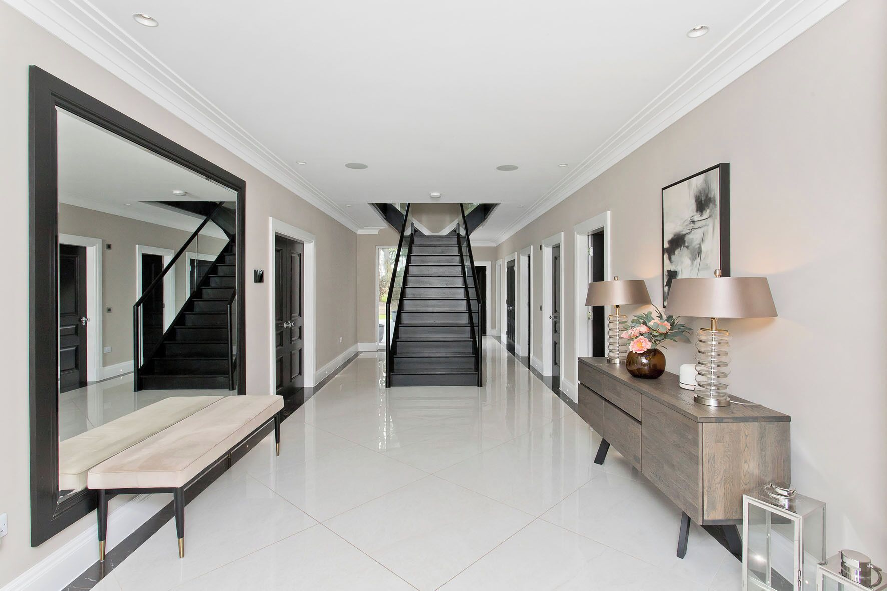 A hallway with a staircase, a bench, a console table with lamps, and a large mirror.