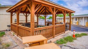 A wooden gazebo with tables and a grill, adjacent to a motel with rooms.