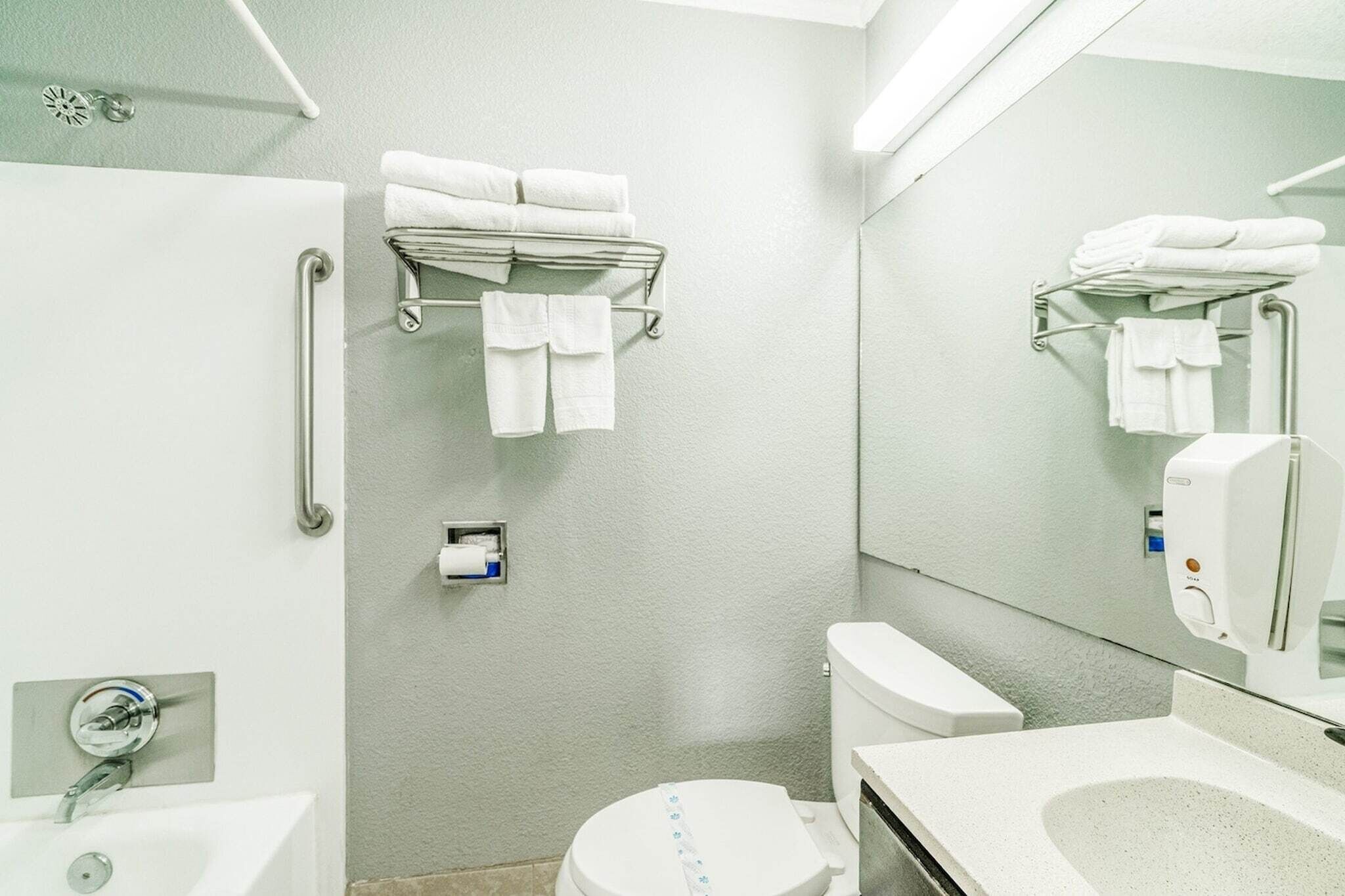 A bathroom with a toilet, sink, handrails, towel rack, and a soap dispenser.