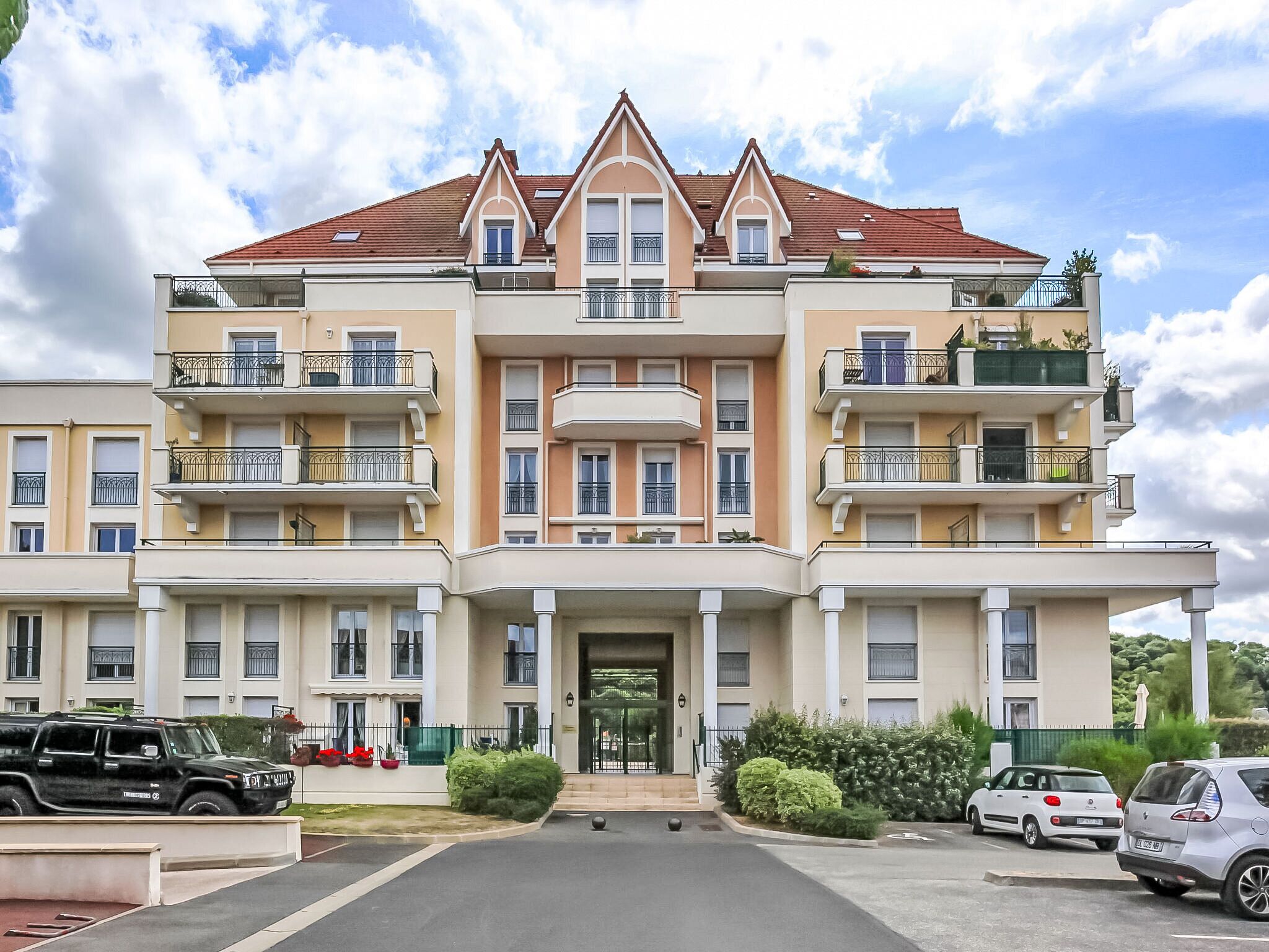 A multi-story residential building with balconies, a central entrance, and a parking area with vehicles.