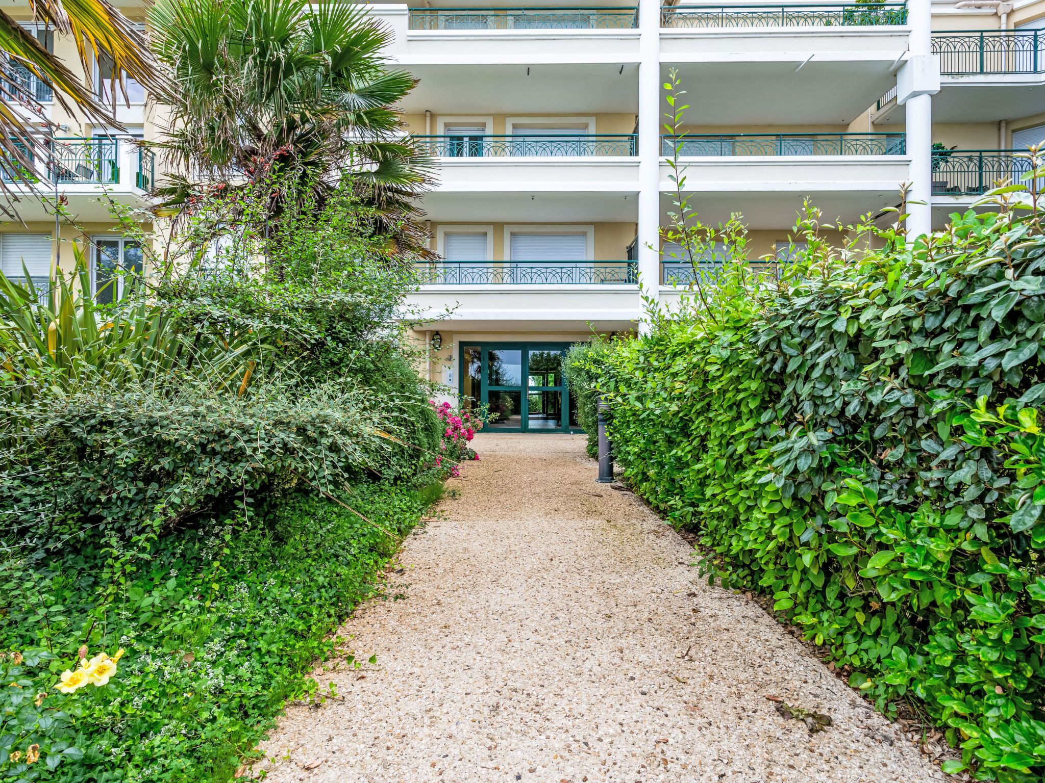 A multi-story hotel with a gravel pathway leading to the entrance, surrounded by lush greenery and a palm tree.