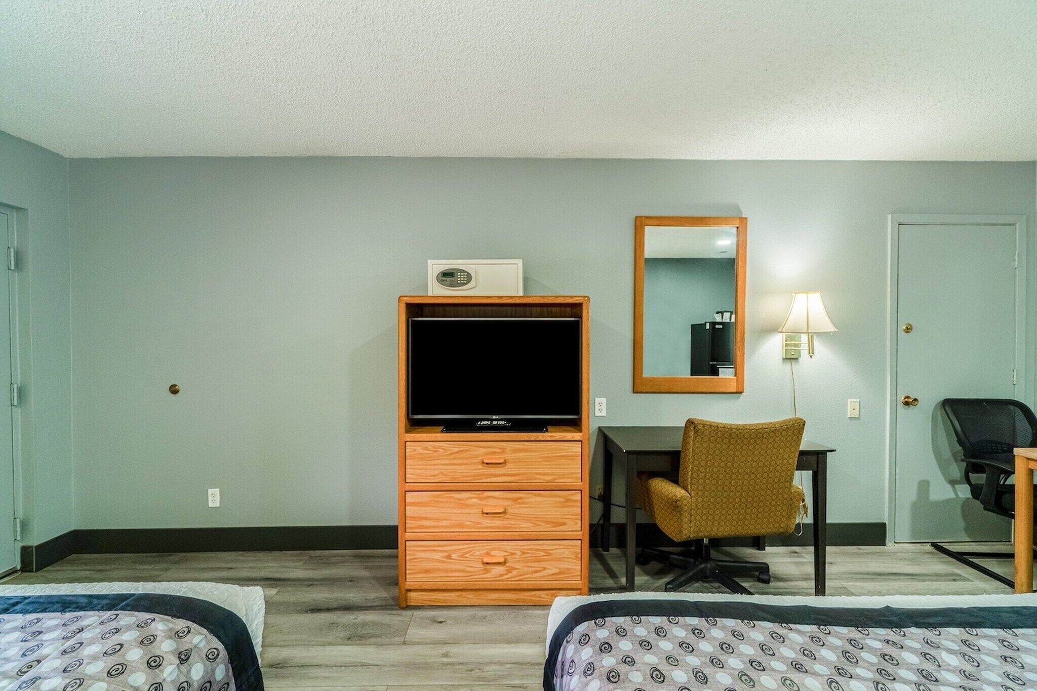 Habitación de hotel con dos camas, un aparador de madera con televisor, un escritorio con silla, un espejo y una lámpara.
